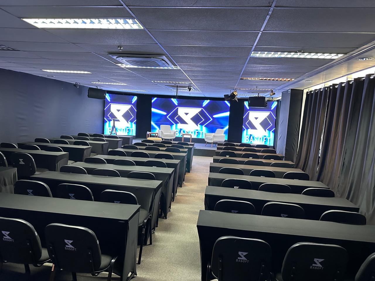 Auditório da BASOFT montado no formato escolar, com mesas e painéis de LED ao fundo